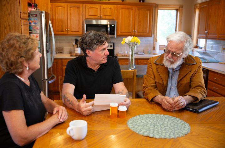 Aric Putnam sitting at a kitchen table with an older couple, prescription bottles on the table between them.