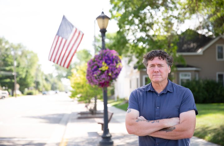 Aric Putnam standing on a tree-lined main street beside a lamppost with an American flag and a hanging flower basket.